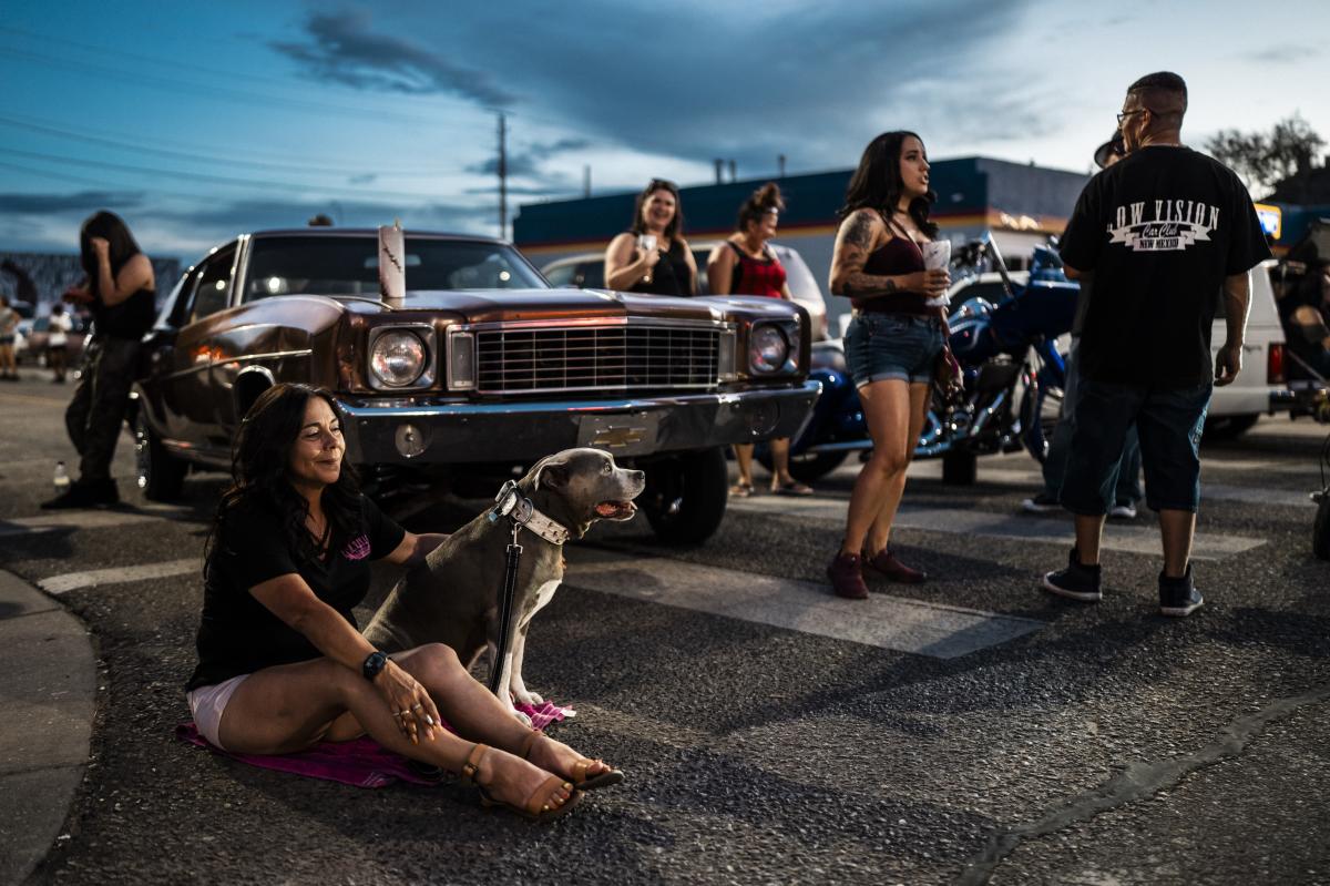 An older dog named Duke sits quietly on a towel among a lively lowrider gathering at Seventh and Central during a warm summer evening.