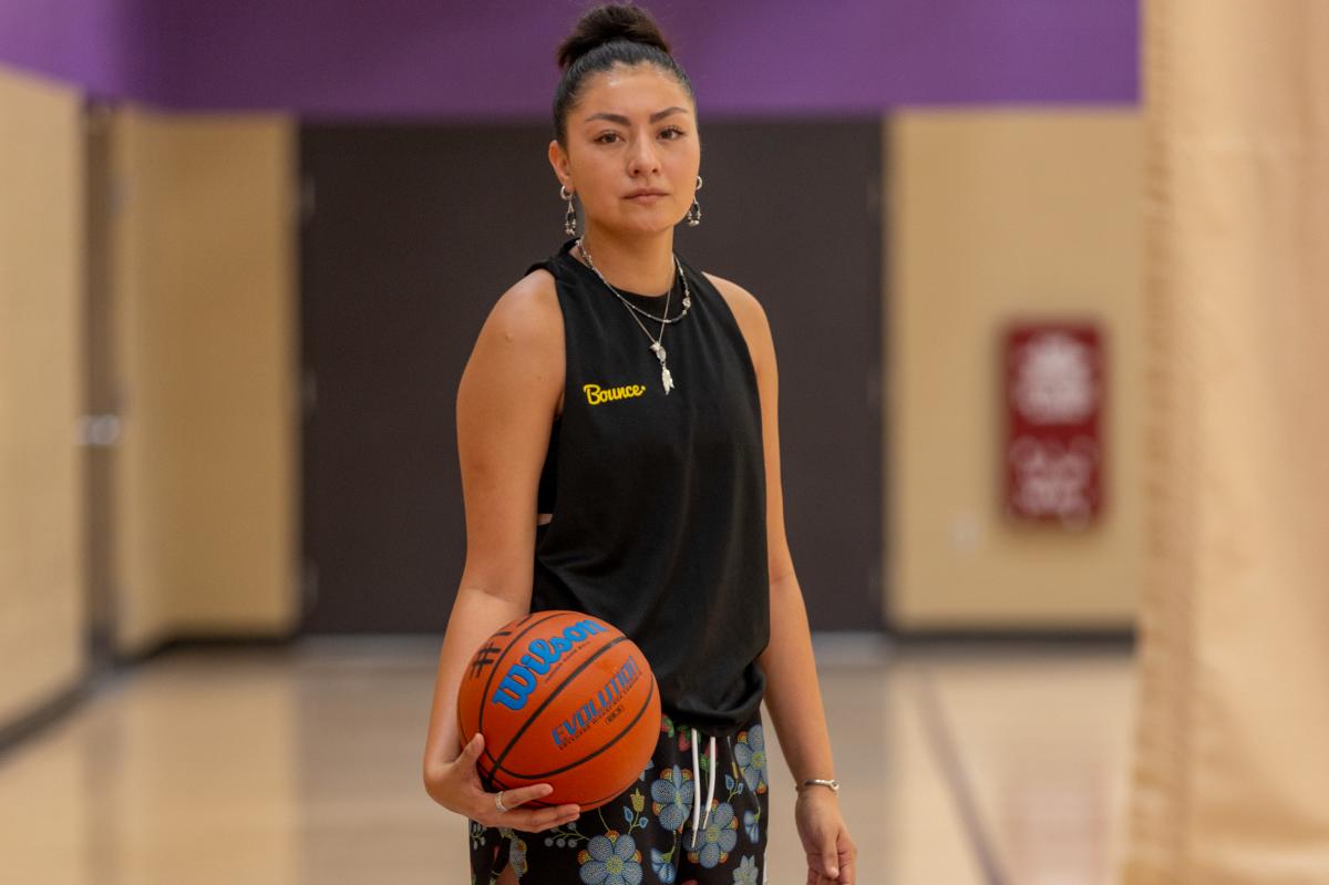 A woman stands on a basketball court holding a ball, wearing a black top, floral shorts, and colorful sneakers.