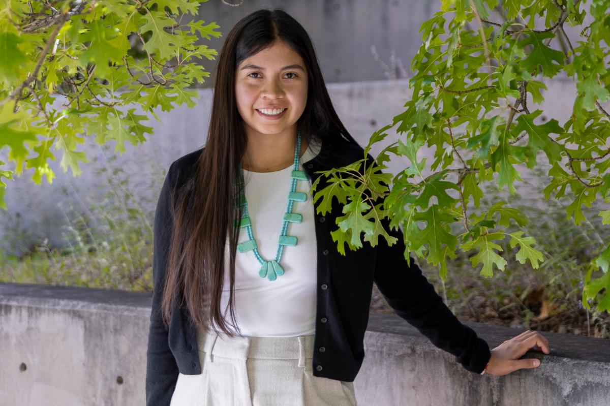 Smiling woman with long hair stands outdoors under lush green leaves, wearing a black cardigan, white top, turquoise necklace, and cream pants.