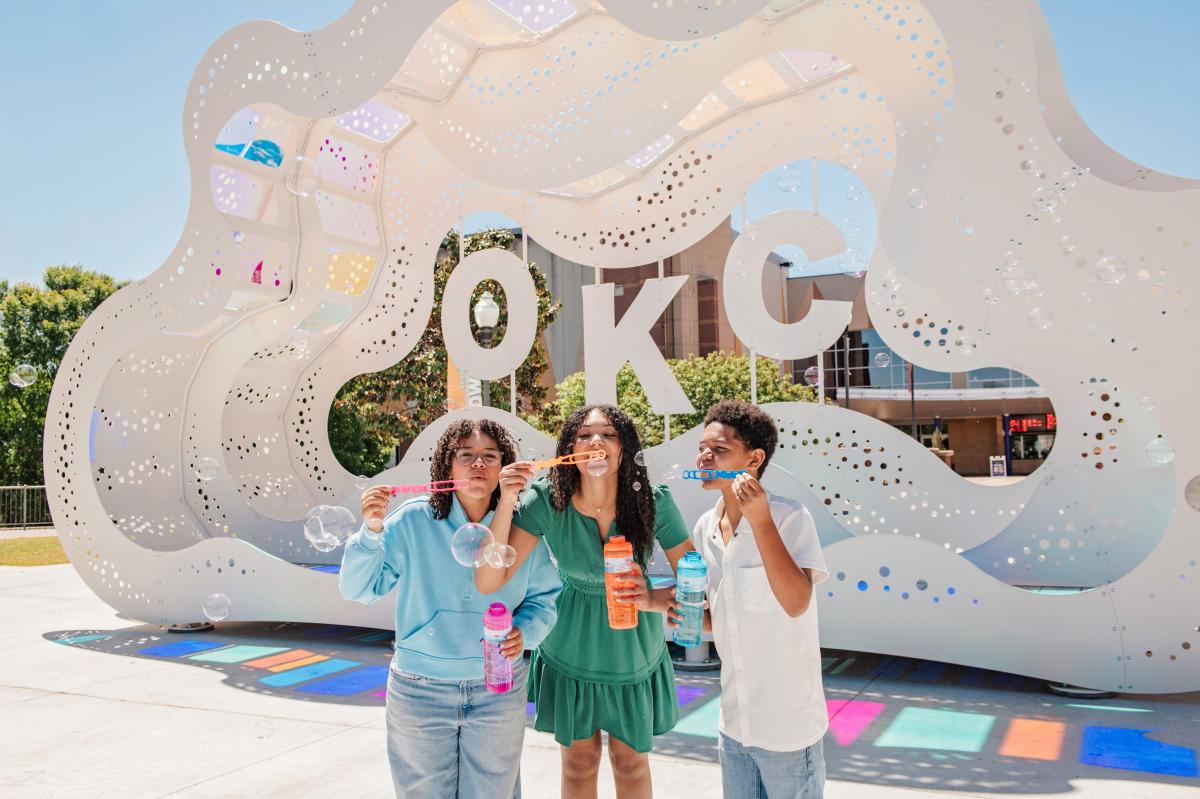 Three children blow bubbles in front of Cloud Embrace art installation in Bricktown