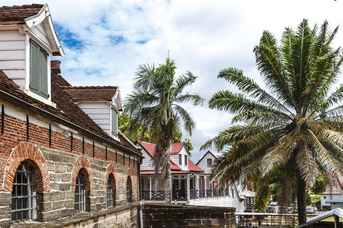 A row of historic buildings with red brick facades and white trim line a street. Palm trees sway in the breeze, and a clear blue sky is visible above.