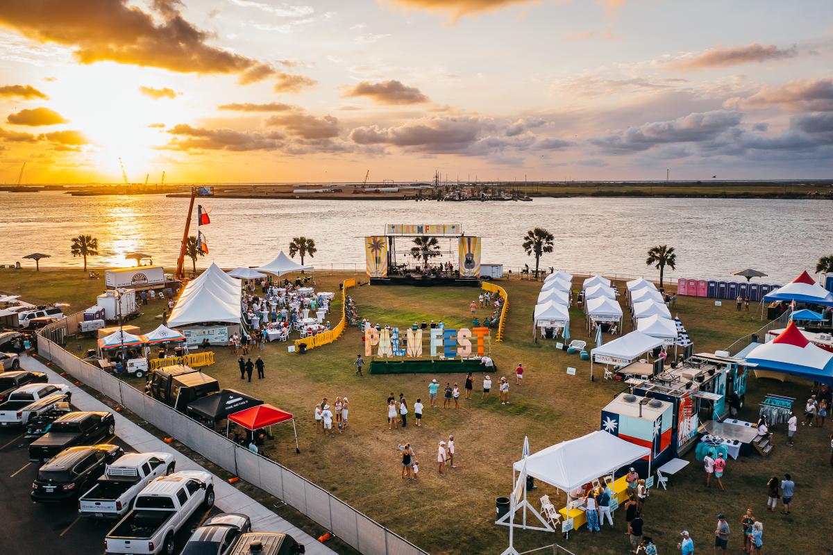 Aerial view of a park right on the ship channel. In the park are tents, a music stage, people, and a large sign reading "PalmFest."