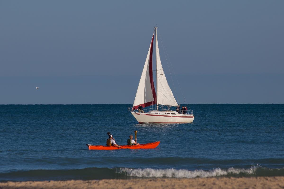 A bright red kayak with two paddlers in it sit in the water. Behind it is a white sailboat. The water is a deep blue with a single white cresting wave near the sandy shore.