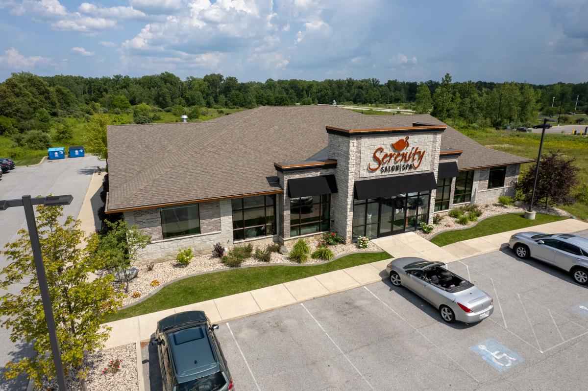 Aerial shot of the Serenity Salon & Spa building with a light stone exterior, black awnings, and a brown-shingled roof. The entrance features large glass doors, with landscaping of bushes, flowers, and stone accents. Two cars are parked out front, including one in a handicapped spot. The building is set against a backdrop of trees and grassy fields beneath a partly cloudy sky.