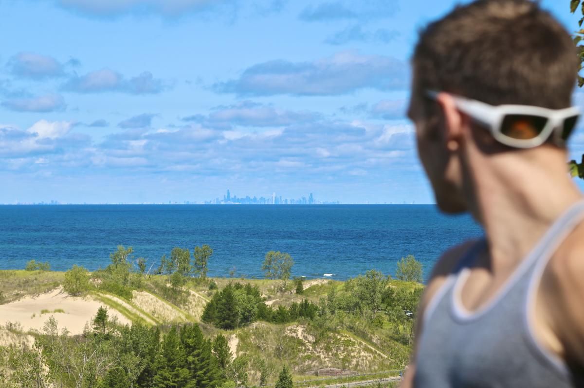 A landscape with vegetation-covered dunes, a blue lake, and the Chicago skyline are in the background. In the foreground is the back head of a man with sunglasses on his head.
