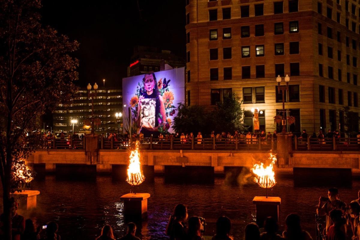 Braziers burn on the river and a mural of a young girl is illuminated in the background.