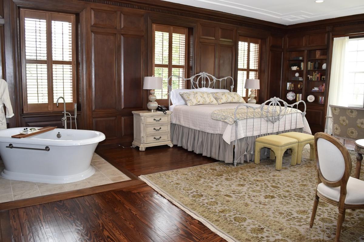a room at an inn, the room has wood paneling, a bathtub, a side-table, a bed with a white comforter, foot stools and a chair with hardwood floors and a white and gold floral rug.