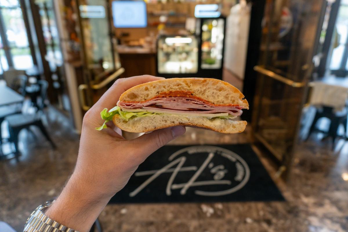 A hand holds a sandwich with layers of lettuce, turkey, and salami on ciabatta bread in a cozy cafe. Background shows a counter and empty tables.