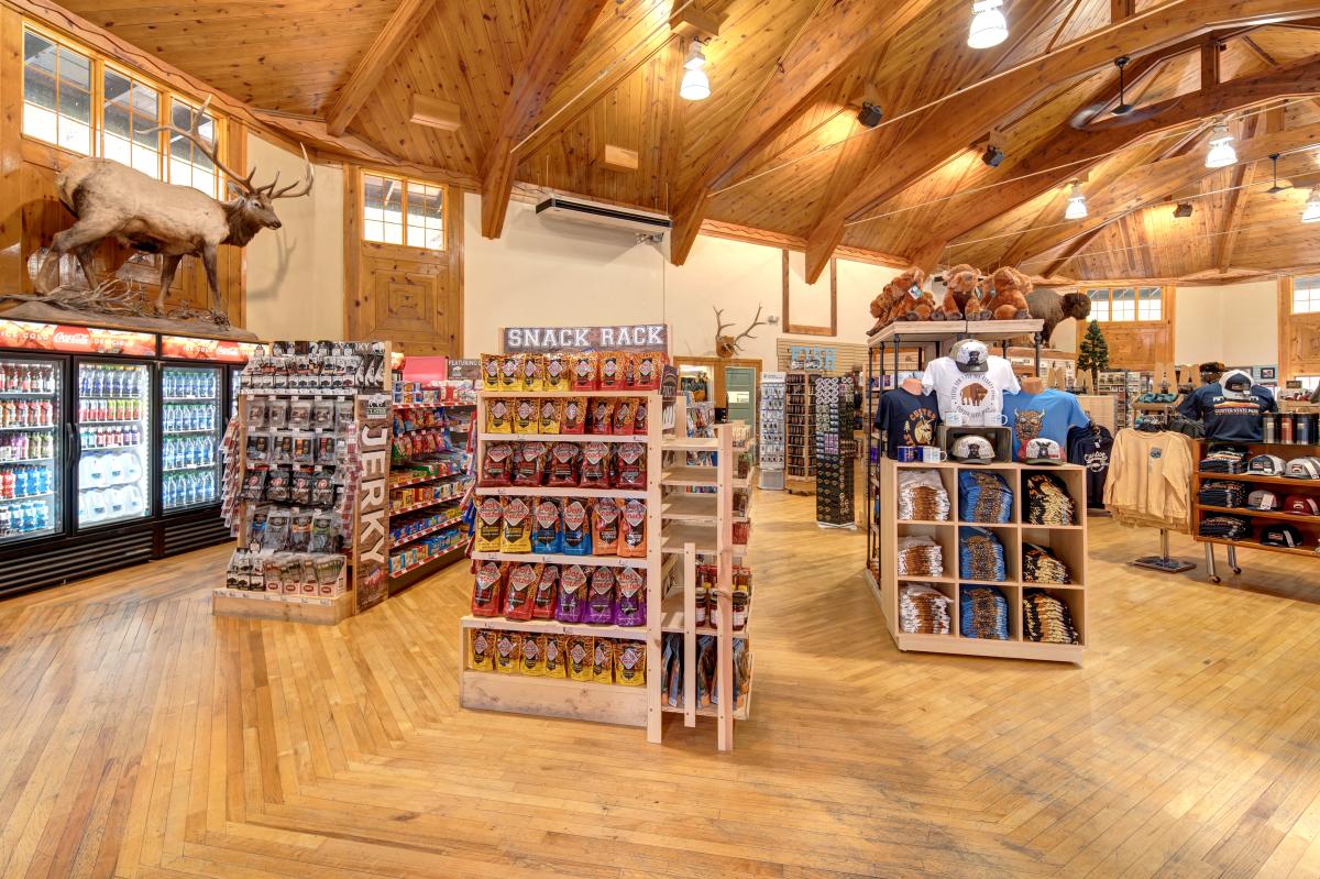 Interior of a rustic lodge-style gift shop with vaulted wooden ceilings, exposed beams, and hardwood floors. Displays of snacks, drinks, souvenirs, hats, and folded apparel fill the space. A large mounted elk stands above refrigerated beverage cases on the left, while stuffed animals and wildlife-themed merchandise are arranged throughout the store. Warm lighting and natural wood finishes create a cozy, outdoorsy atmosphere.