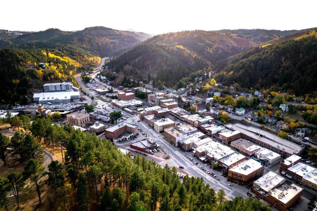 Aerial view of downtown Deadwood, South Dakota, surrounded by pine-covered Black Hills with fall foliage glowing across the hillsides.