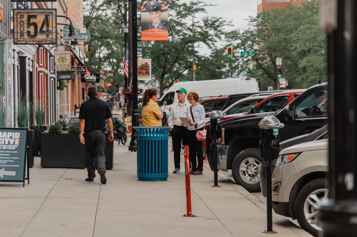 Downtown Rapid City Ambassador in a yellow shirt speaks with visitors on a busy sidewalk lined with storefronts, signs, parked cars, and street banners.