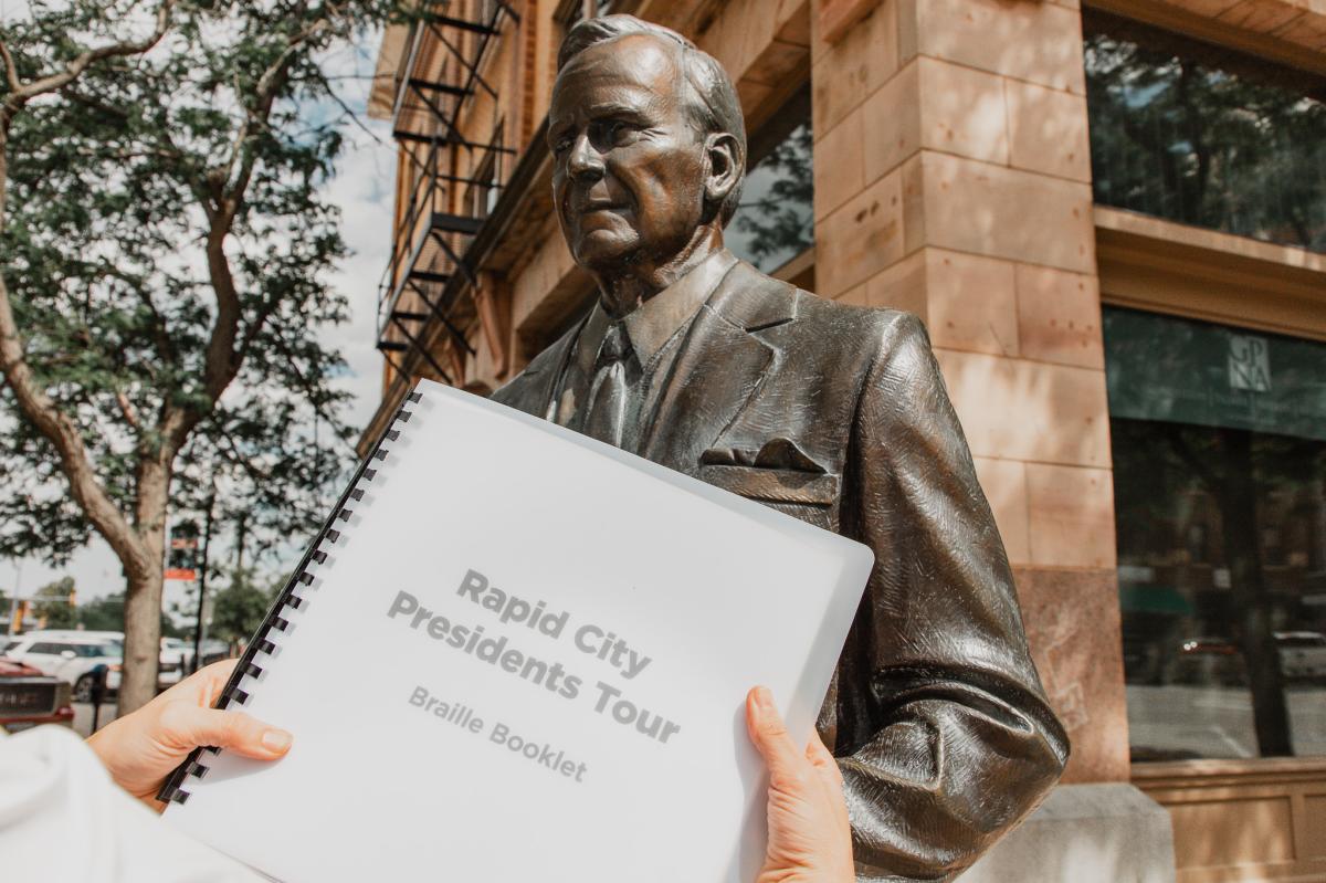 A woman holds up the Rapid City Presidents Tour braille booklet in front of a bronze president statue