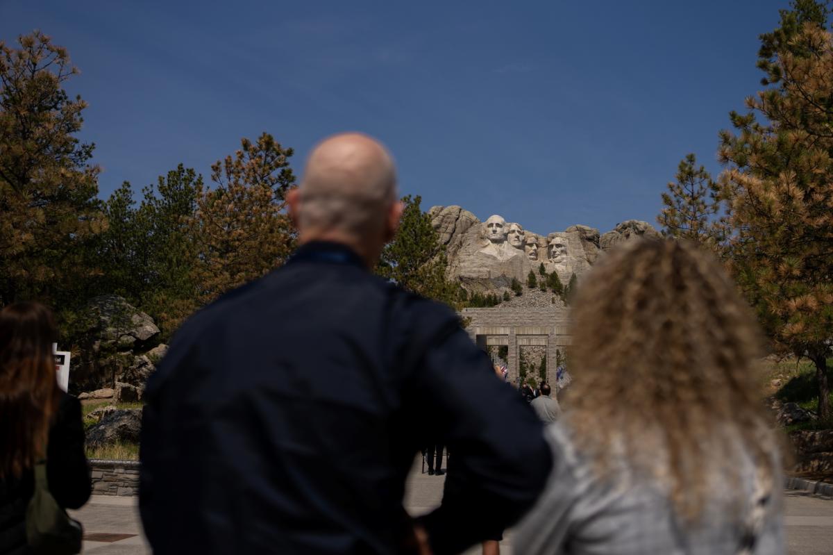 A man and a woman, slightly out of focus in the foreground, look up at Mount Rushmore, in focus in the distance positioned between their heads.