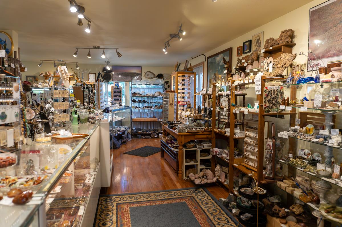Cozy shop filled with various rocks, minerals, and trinkets on wooden shelves and glass displays. Warm lighting and hardwood floors create an inviting atmosphere.