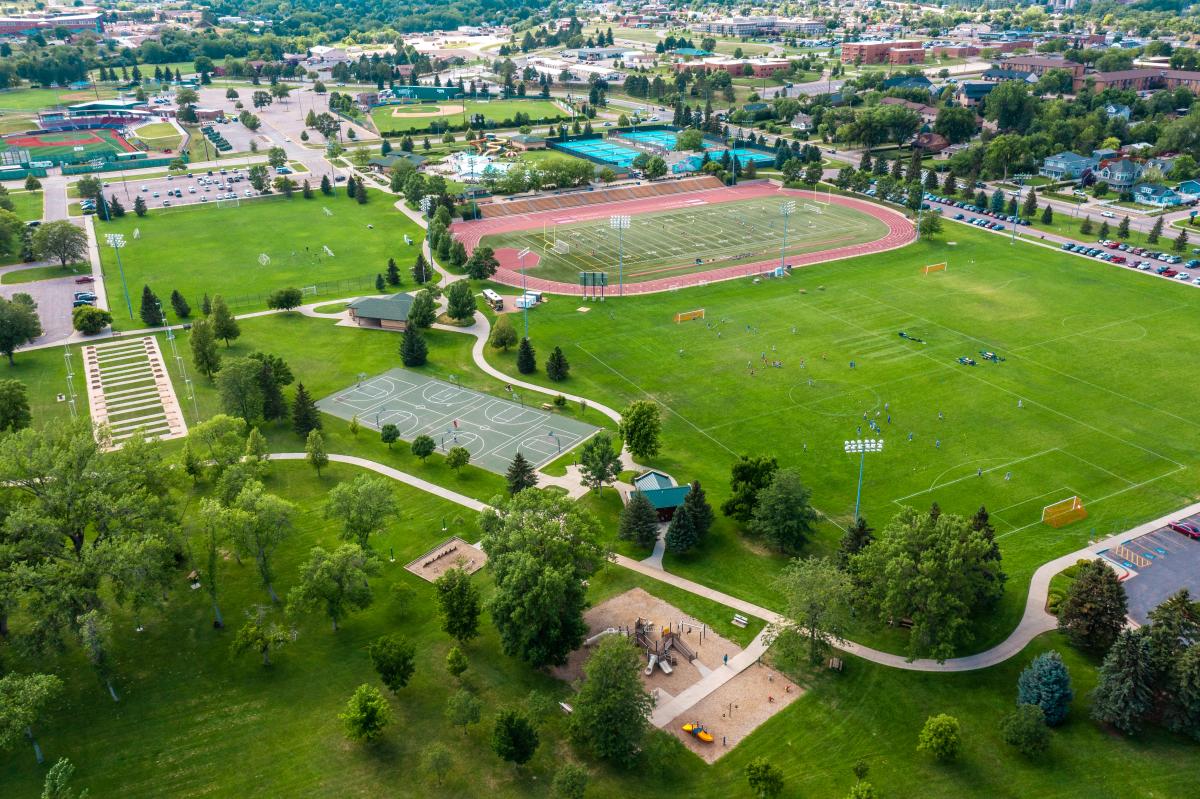 Birds eye view of soccer field, playground, horseshoe, track, basketball court, and pool surrounded by housing.