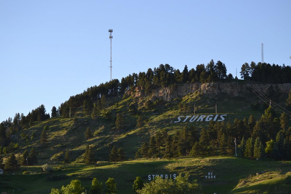 Green, pine-covered hillside in Sturgis, South Dakota, with large white “STURGIS” letters visible below rocky cliffs beneath a clear blue sky.