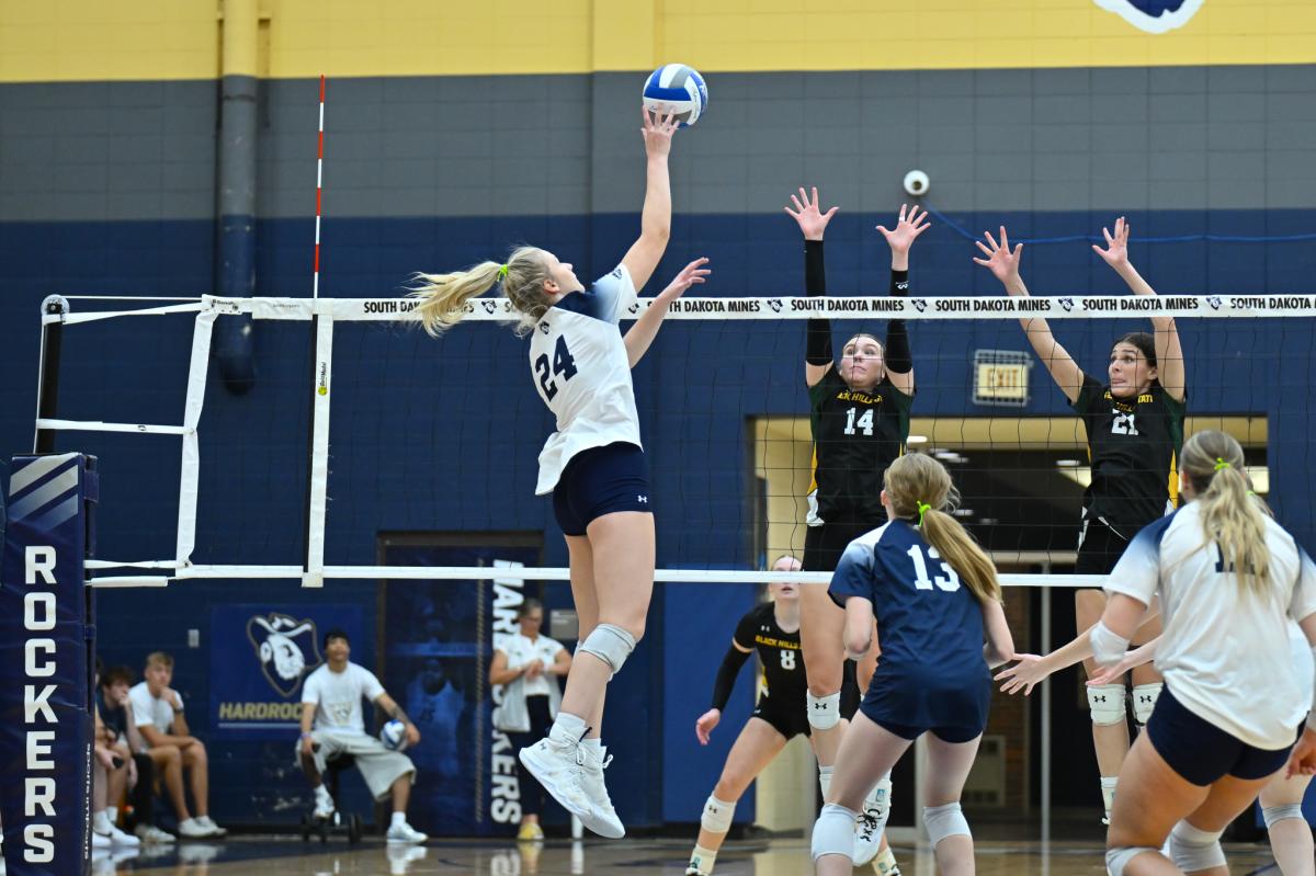 A volleyball player in a white jersey leaps to tap the ball over the net above two defenders in black jerseys.
