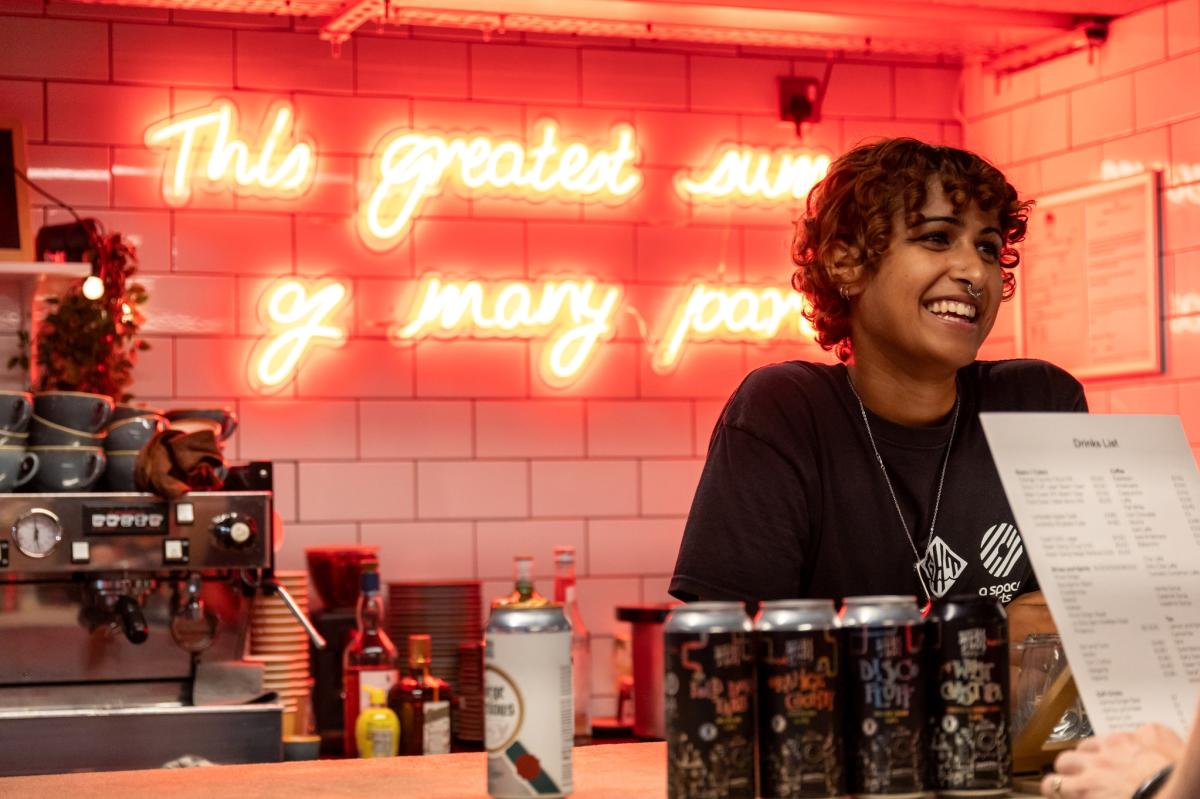 Woman smiling behind a cafe counter at God' House Towe as customer looks at menu