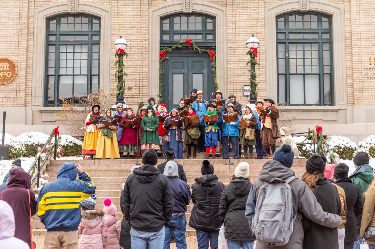 Carolers in vintage winter attire singing on snowy St. Charles stage with festive Christmas decorations.