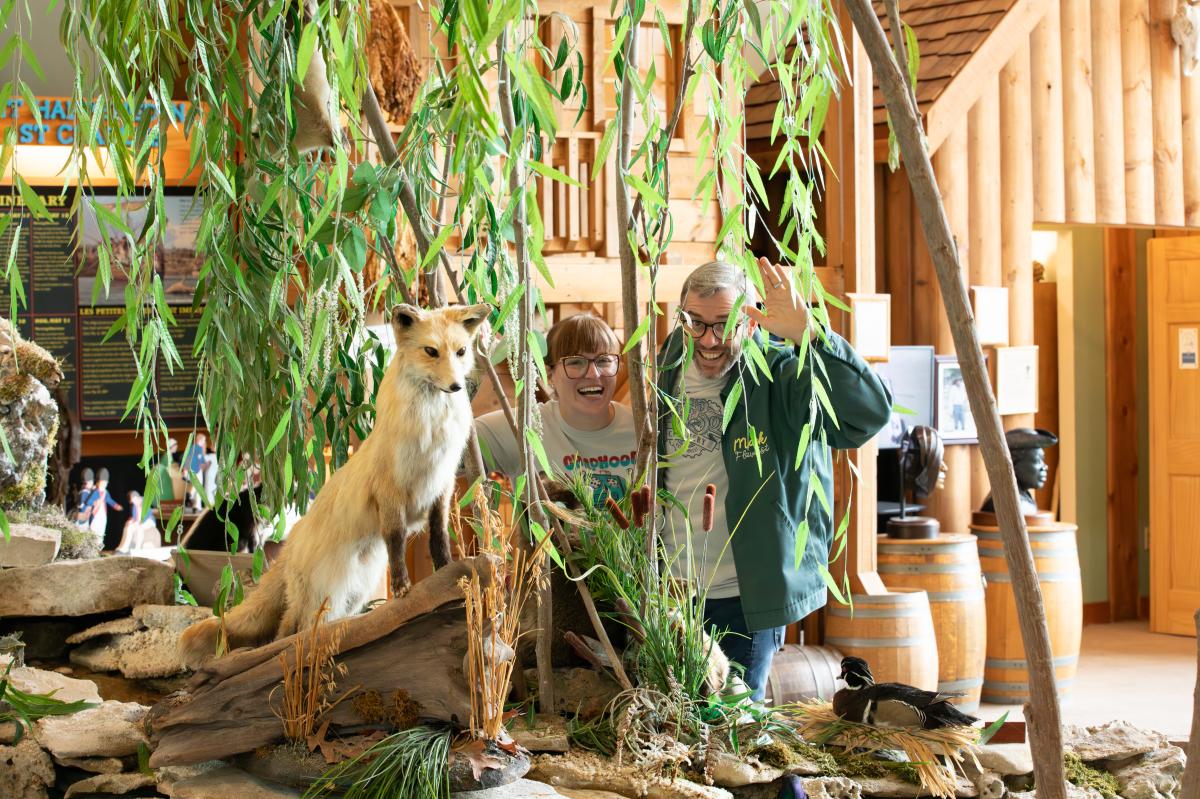 couple standing behind display of stuffed fox and fake tree in museum, making funny faces.