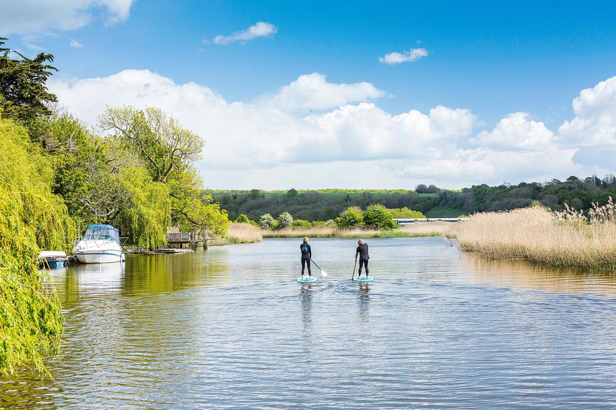 Two paddle boarders on the River Arun, with a boat moored to the left and grassy bank to the right, in Sussex