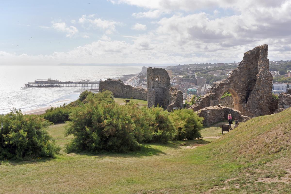 A view of the ruins of Hastings Castle sat high on a hill with the town of Hastings, the pier and sea in the distance