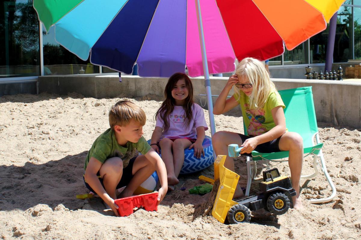 three children playing in sand under rainbow umbrella