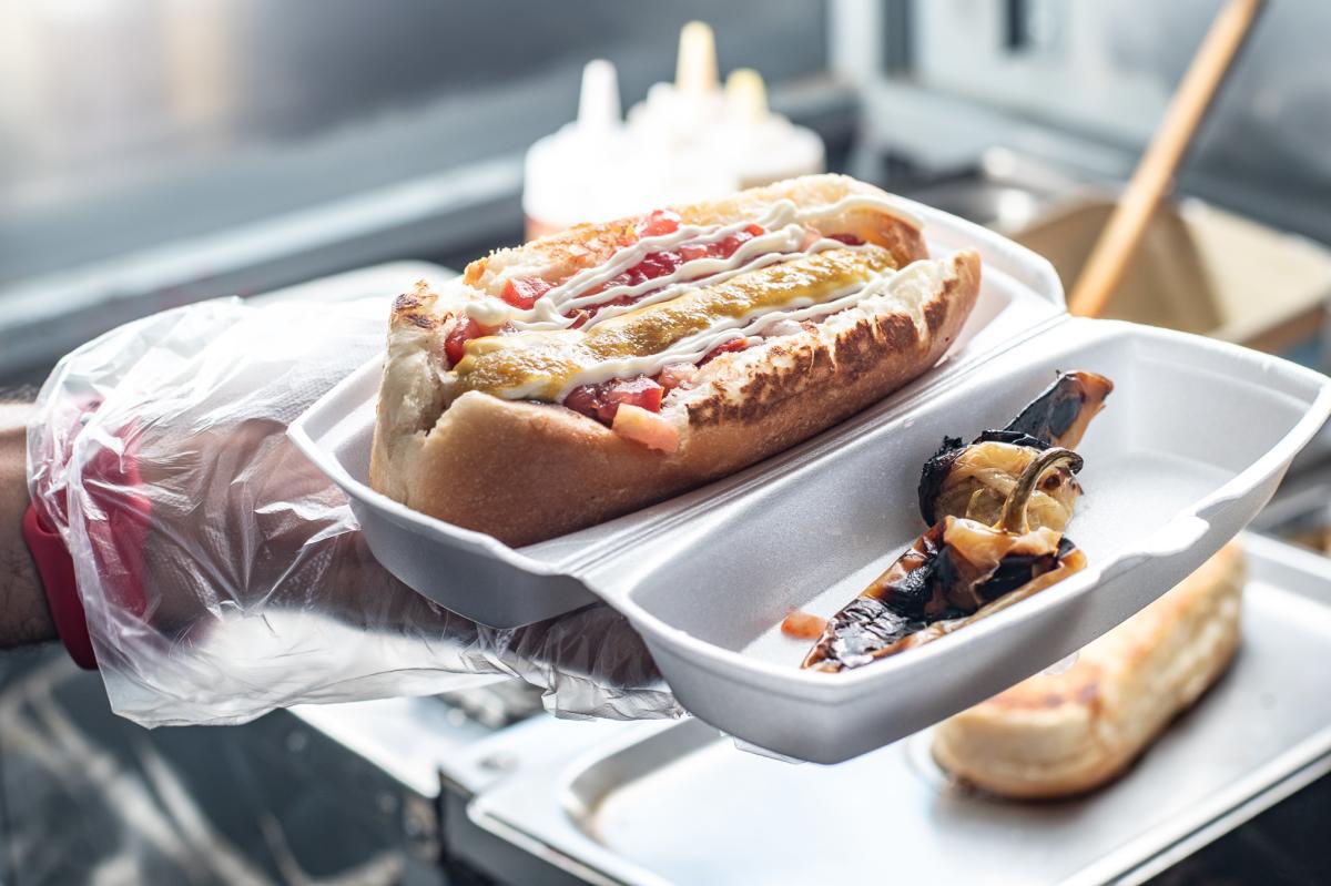 person holding a takeout container with a sonoran dog and peppers