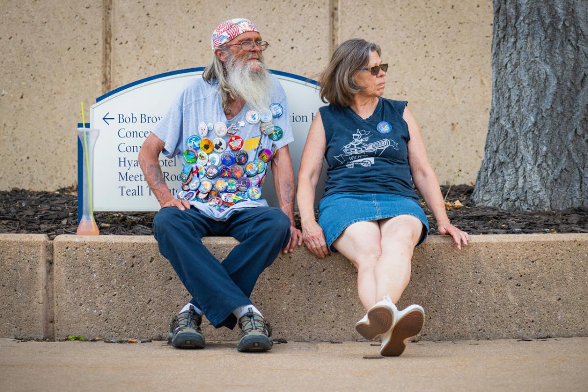 A man sits on a curb dressed in a T-shirt covered in Riverfest buttons from past festivals during the Riverfest in Wichita.