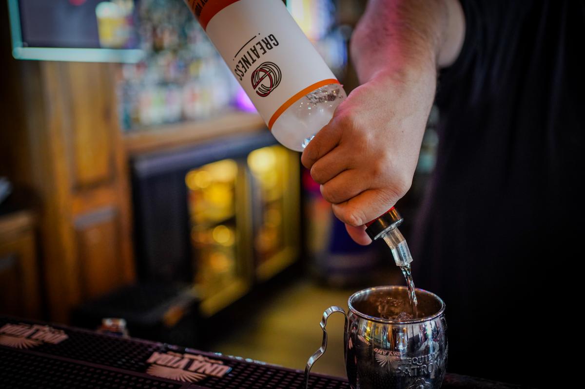 A hand pours Greatness Vodka from a bottle into a glass at a bar