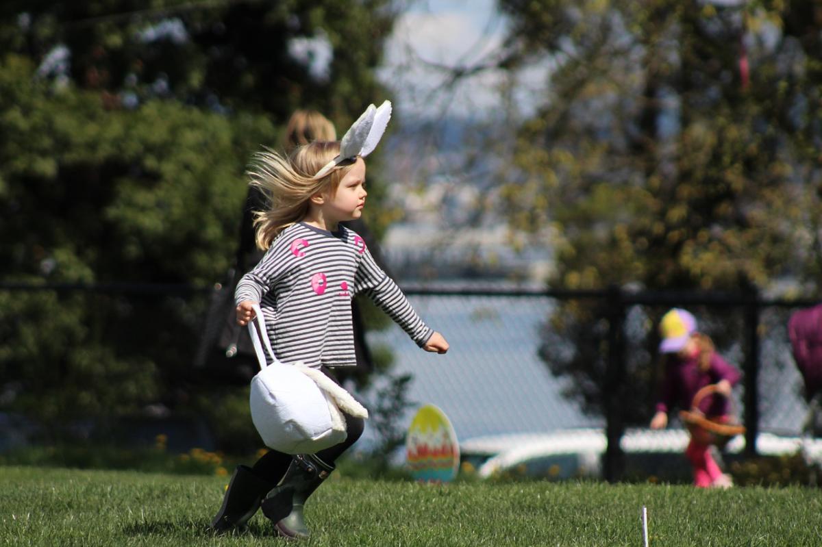 A young girl runs through a yard looking for Easter eggs with a backet in hand and bunny ears on her head