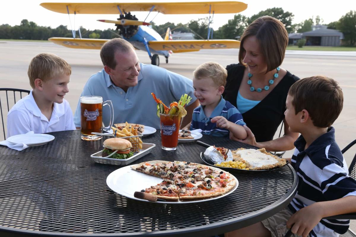 A family enjoys burgers and pizza on the patio at Stearman Field while a small prop plane is parked on the landing strip behind them