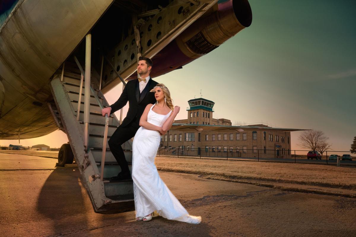 A bride and groom pose on the steps of an airplane on the tarmac at Kansas Aviation Museum
