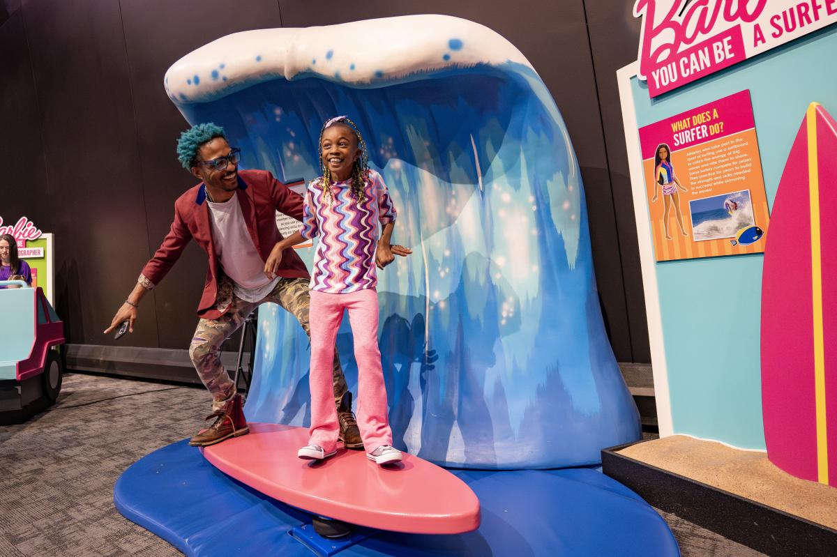 A young girl enjoys a surf board display at the Barbie You Can Be Anything exhibit