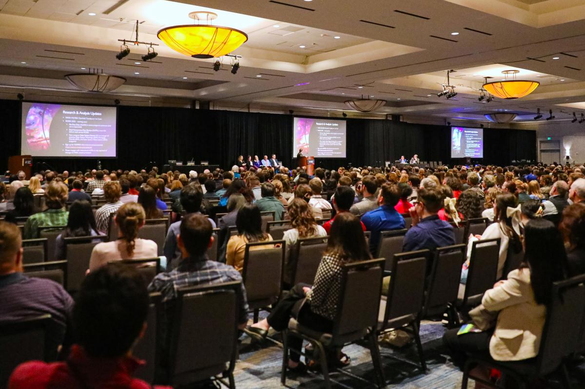 The Lunar Planetary Science Conference in The Woodlands Texas. A large ballroom is packed full of students and researchers of astronomy. A panel of five experts and a moderator are on the stage. Three projector screens read, "Research & Analysis Updates."