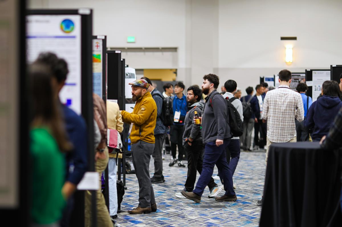 The Lunar Planetary Science Conference in The Woodlands Texas. A large ballroom is packed full of students and researchers of astronomy looking at each other's poster presentations. The central figure is a man in a tan jacket and checkered newsboy cap.