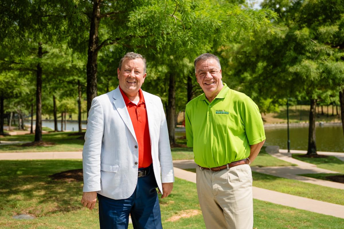 Two men smile at the camera. On the left is Nick Wolda, President of Visit The Woodlands, wearing blue jeans, a bright red polo, and a white blazer. On the right is Chris Nunes, wearing a lime green polo and khaki slacks with a brown belt. They are standing in a park with bright green grass, bright green trees, and a branch of The Woodlands Waterway.