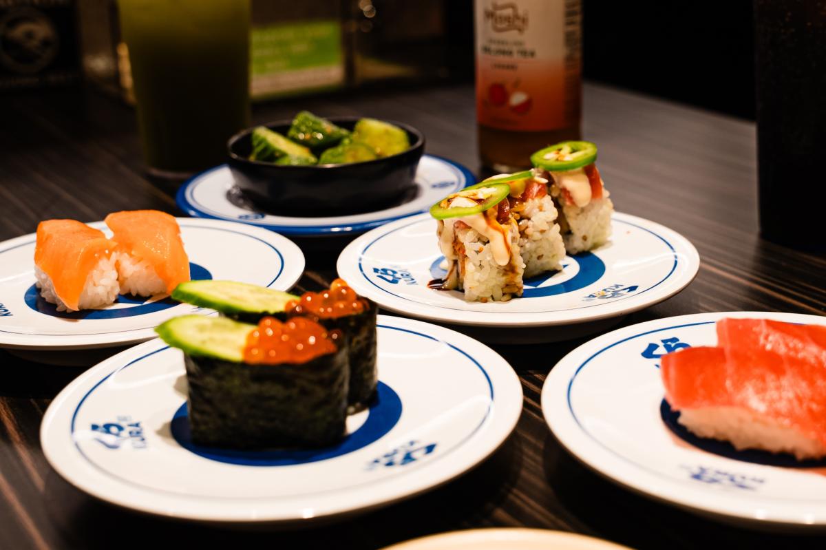A spread of 5+ single-serve plates of Japanese cuisine: a bowl of seasoned brussels sprouts; one plate of sushi topped with a creamy sauce and jalapeno slices; two types of nigiri; and one dish that's wrapped in seaweed and topped with cucumber slices and orange Tobiko.