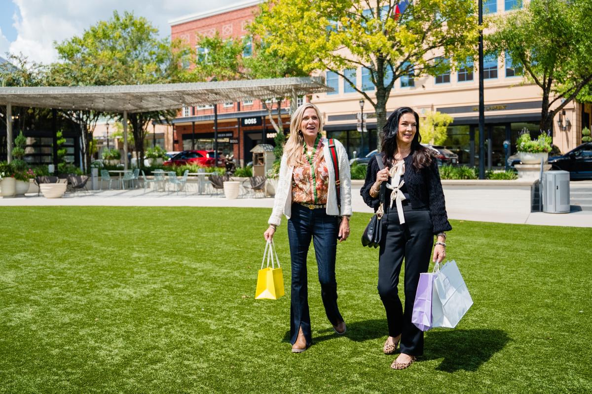 Two women dressed for fall walk through Central Park at Market Street. The blonde woman (left) is carrying a small yellow shopping bag from Kendra Scott. The dark-haired woman (right) is carrying larger bags in purple (Hemline) and white (Golden Gray Boutique).