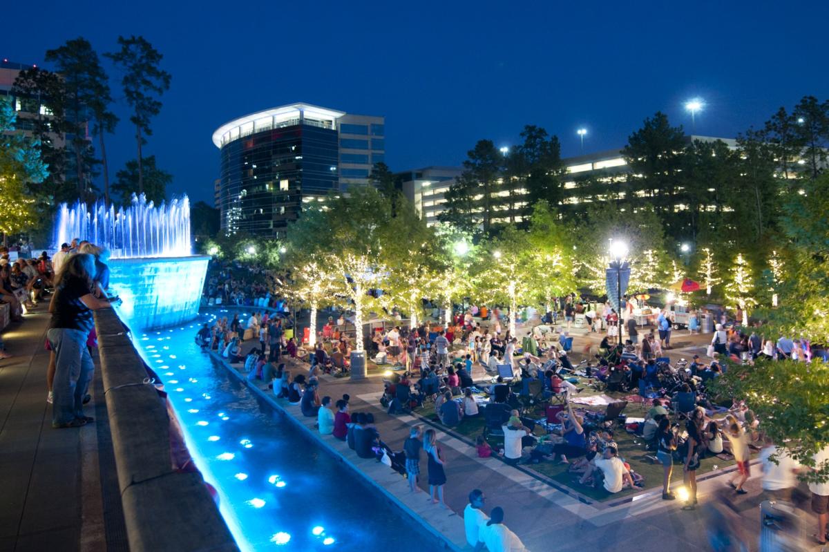 Waterway Nights at Waterway Square. The photo is taken from an upper level of the plaza, overlooking The Fountains at Waterway Square (glowing blue) and a crowd of live music fans. The trees are wrapped in golden fairy lights.