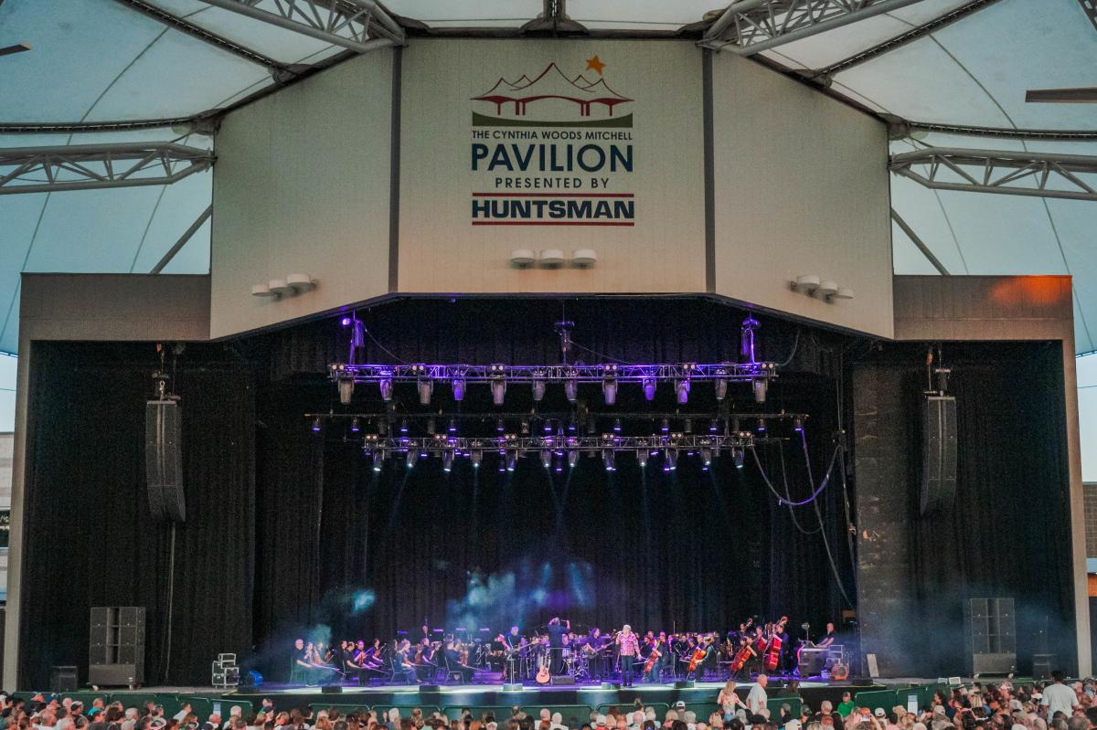 A full orchestra plays on The Cynthia Woods Mitchell Pavilion main stage. The viewer is sitting in the center of the crowd, directly facing the stage. The area is set with light fog and purple spotlights.
