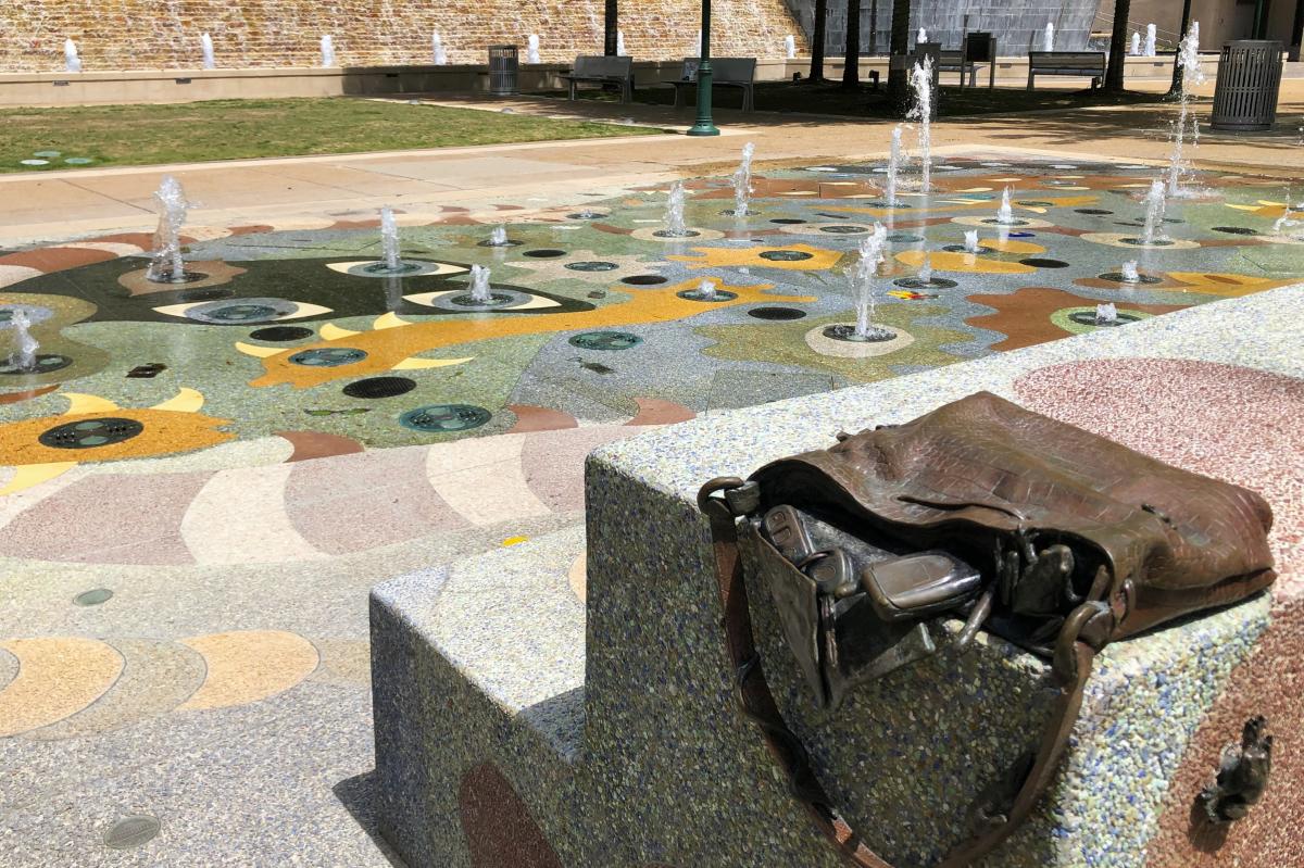 A public art sculpture shaped like an old crocodile skin purse, which is falling open over the side of a concrete bench. Contents include a flip phone, car keys, a pen, and two large frogs. The art piece continues on the ground behind it: a splash pad depicting simple organic shapes and fantasy creatures with eyes that spout water.