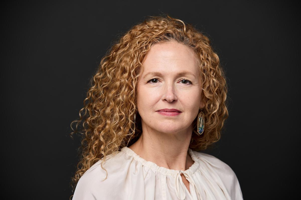 A professional headshot of Kim Lowe, who has curly red hair and is wearing a white blouse.