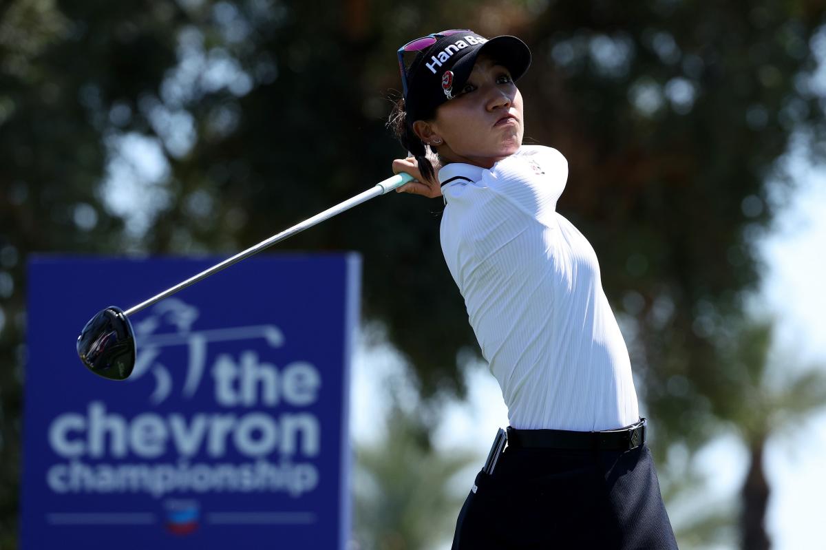 A young South Korean woman (playing for New Zealand) swings her golf club. She is wearing a white long-sleeved athletic shirt, dark pants, and a black hat with pink sunglasses rested on top. Behind her is a blue sign with the Chevron logo and white letters that read, "the chevron championship."