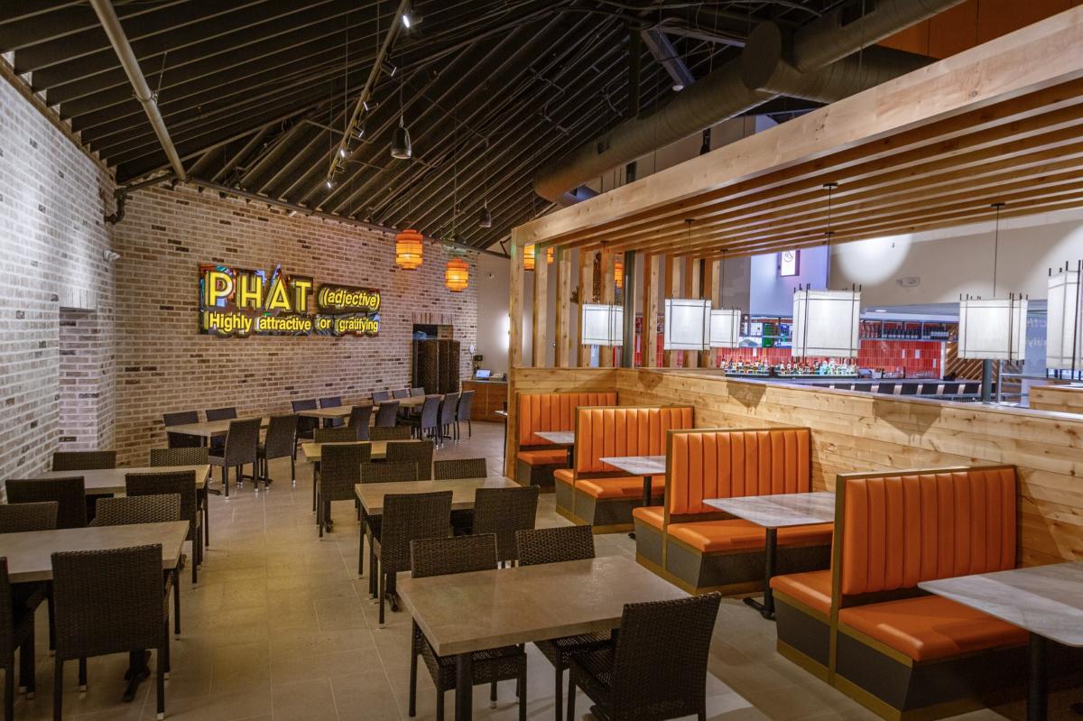 The camera faces the interior of the Phat Eatery Woodlands location. There are brick walls, sloped metal ceilings, and tan-tiled floors. There are eight tables visible, each with four wicker chairs, along the wall or in a row beside them. In the middle of the space is a slatted wood awning over a row of four bright orange booths, each seating four. Behind the booths is an obscured view of the kitchen. On the brick wall is a yellow neon sign that reads, "PHAT (adjective) Highly attractive or gratifying."