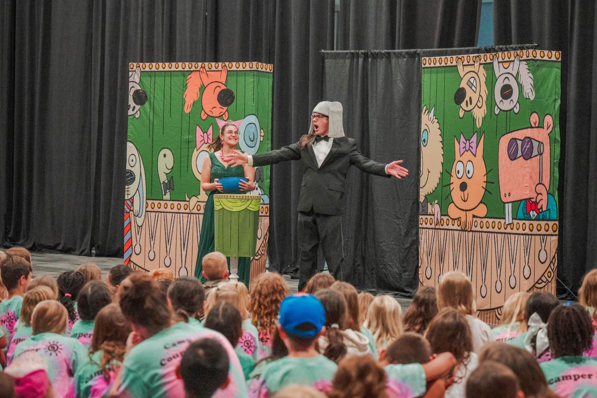 Two actors perform for children at The Pavilion. One actor is wearing a formal suit and an elephant hat, and he sings dramatically. An actress beside him wears a bright green formal gown and tiara. The backdrop depicts cartoon animals. Kids in the audience are wearing matching tie-dye shirts.