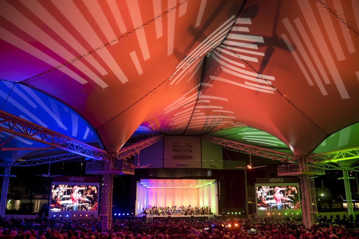 A photo of The Pavilion tents and stage from the crowd. There appears to be an orchestra on stage; the stands are packed with listeners. The tents are illuminated blue, red, and green with striped patterns.