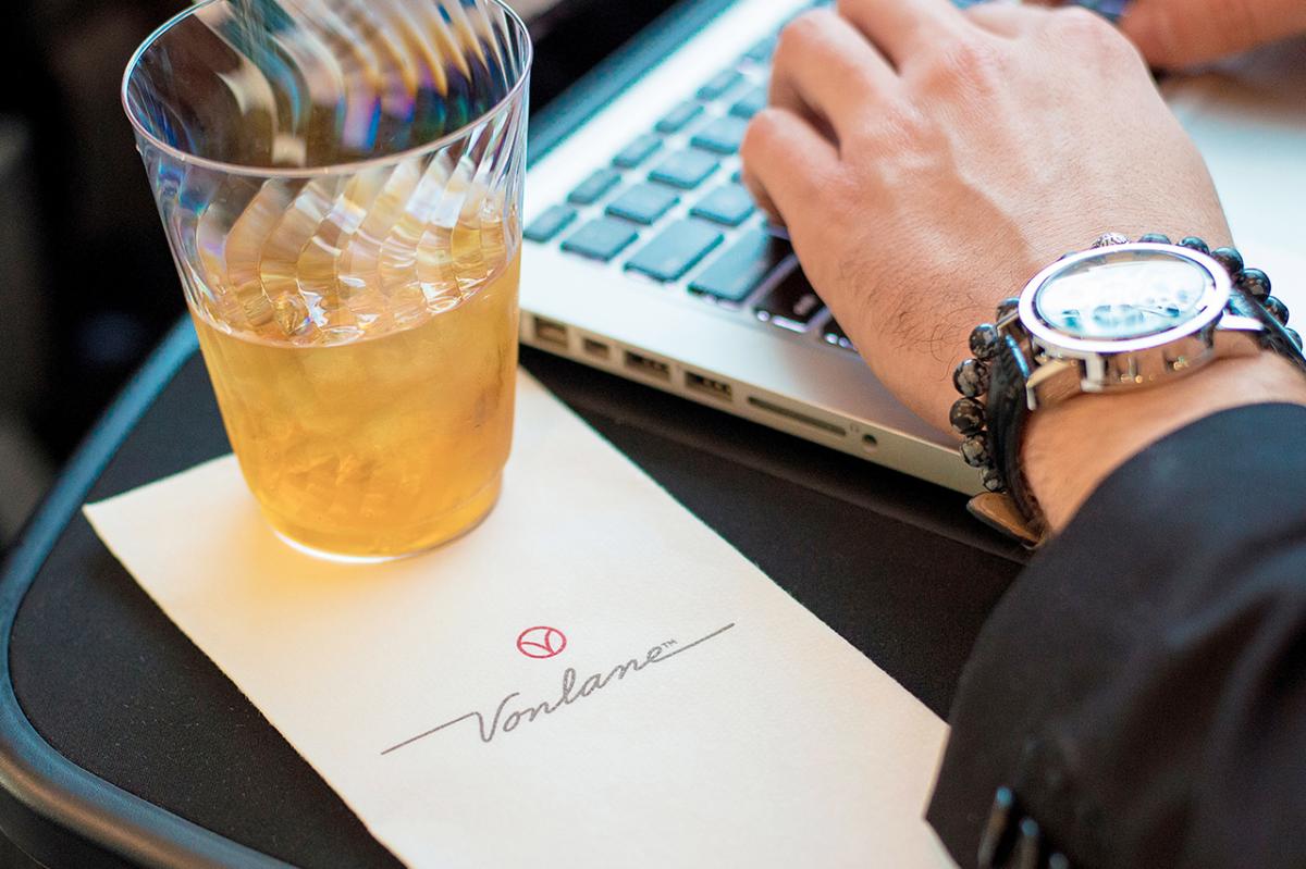 A close-up photo of a Vonlane bus's passenger workstation. A glass of an iced orange beverage sits on a napkin emblazoned with the Vonlane name and logo. Beside it is a man's hand typing on a laptop.
