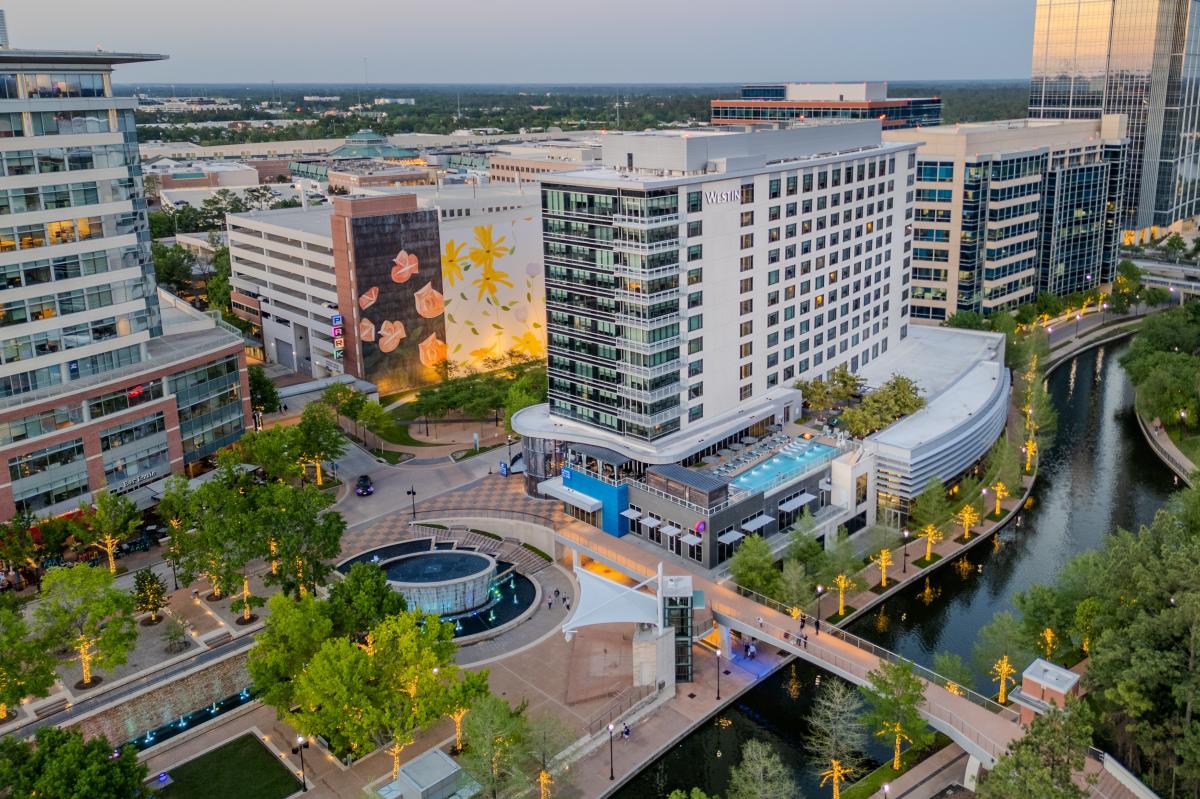 A drone shot of Waterway Square with particular focus on The Westin, a long, white building with even rows of blue-tinted windows. There is a flower mural on the building behind it, The Waterway stretches from the viewer toward The Woodlands Towers in the background.