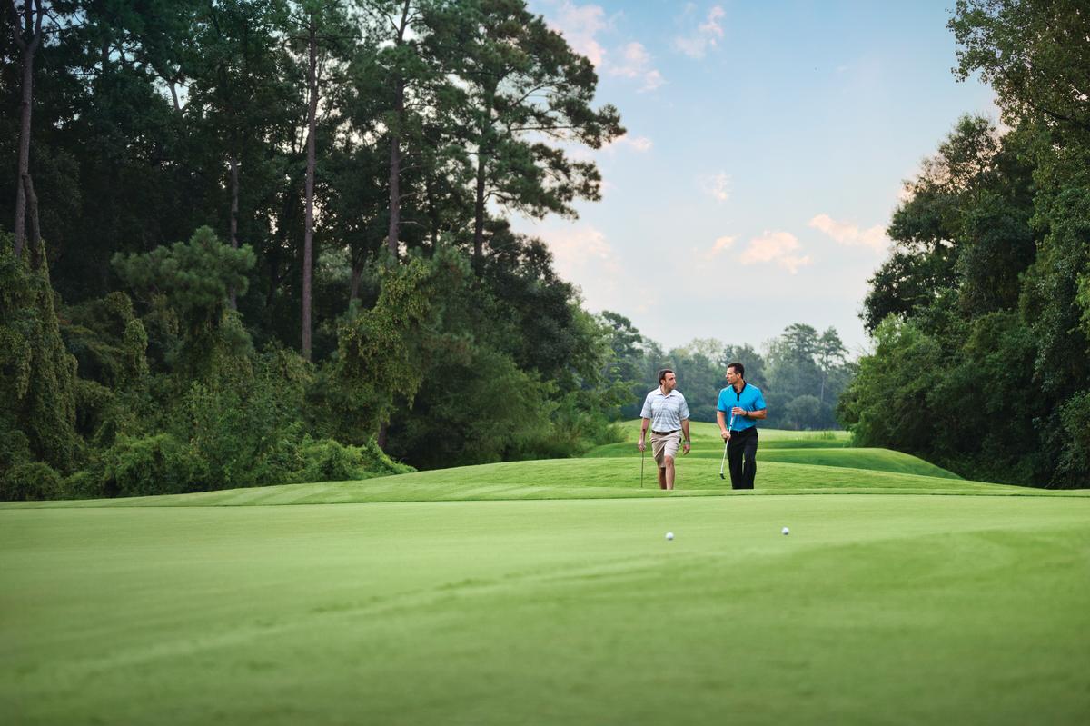 Two men walk across a golf course toward the camera. A lush treeline hugs the course on both sides and wraps around the back, highlighting the light green grass and early morning sky.